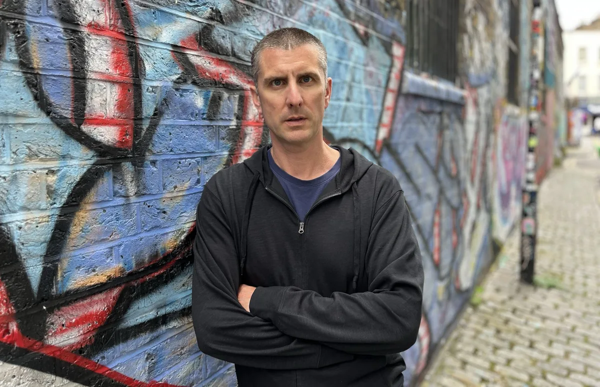 Hugo Bone, arms folded, in front of a graffiti wall in Shoreditch, London